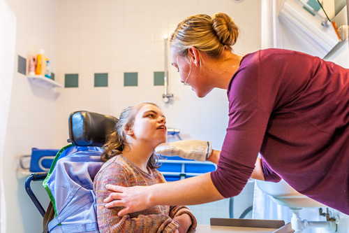 A young woman in a wheelchair looks up as a nurse washes her face.
