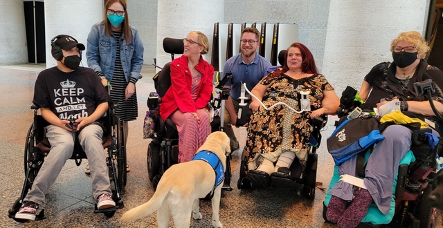 Group of six advocates at the Statehouse, posed together in a line, four with wheelchairs and two kneeling down, along with a service dog