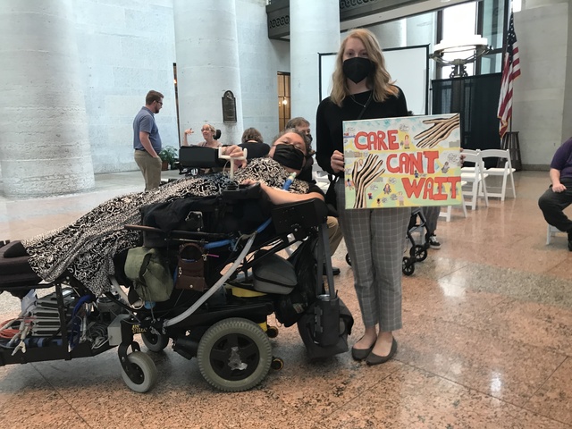 Maria Matzik, in a reclining wheelchair, and Alexia Kemerling, standing and holding a sign of Alicia Hopkin's artwork, which says 'Care Can't Wait' in red letters