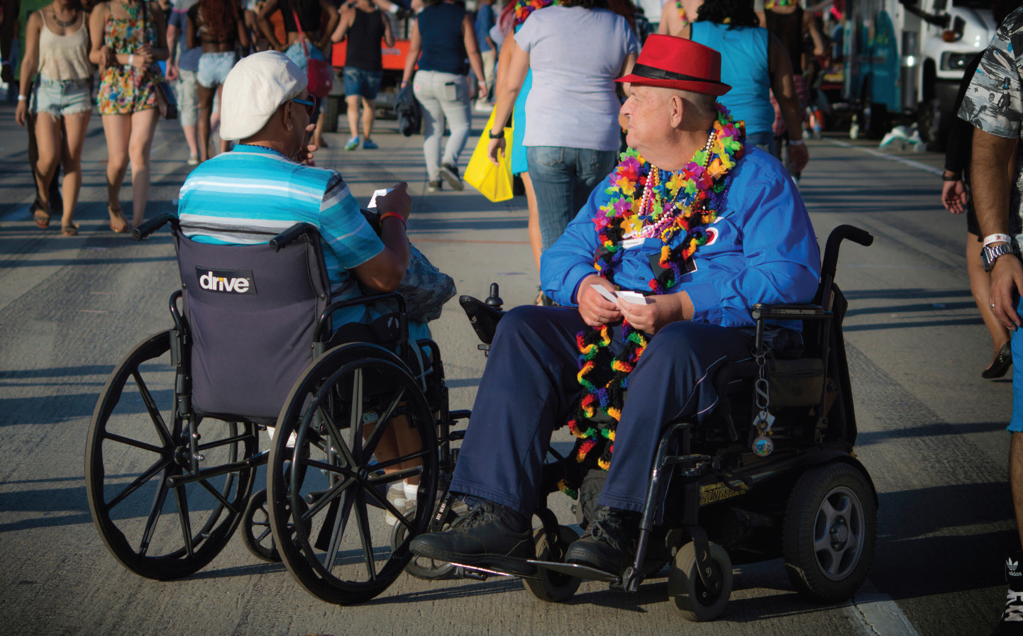 Two men in wheelchairs and fancy hats next to each other in the middle of a busy pathway.