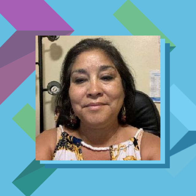 Photo of Norma Ramos-Prater, a Latina woman with light brown skin and shoulder-length black hair. She is wearing a colorful blouse and gold-colored earrings. She is smiling softly at the camera while sitting inside.