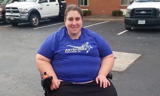 Alicia smiles as she sits in her wheelchair in a parking lot wearing a blue shirt that says ArtSparks