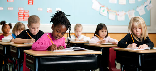 Children sit at their desks in a classroom working on an assignment with pencils and paper