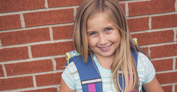A girl with long hair smiles while wearing a blue backpack and standing in front of a brick wall