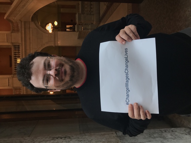 Jason Harris, with rectangle glasses, a black shirt, and curly black hair, holds a sign that says '#ChangeWagesChangeLives'