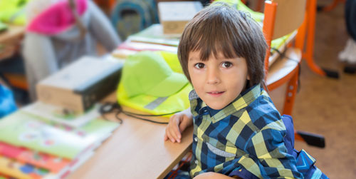A little boy with floppy brown hair sits at his desk in his classroom