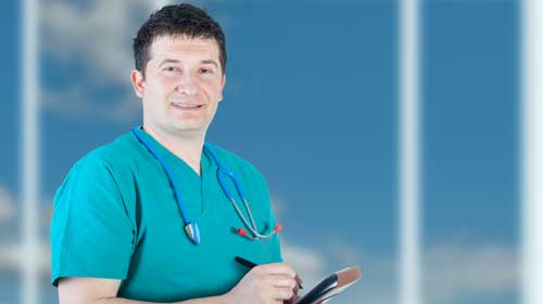 A male nurse wearing scrubs and a stethoscope stands in front of some windows holding a notebook