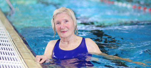 An older woman wearing a blue swimsuit holds onto the wall in a swimming pool