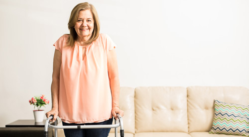 A white woman with shoulder-length brown hair stands in front of a couch facing the camera holding onto a walker and smiling