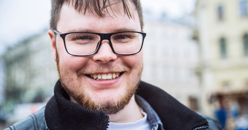 A young white man with short brown hair and a beard smiles at the camera with rain on his glasses