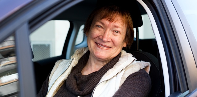 An older woman looks out of the drivers seat window of a car and smiles