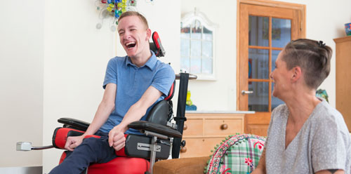 A young man in a wheelchair laughs with his caregiver
