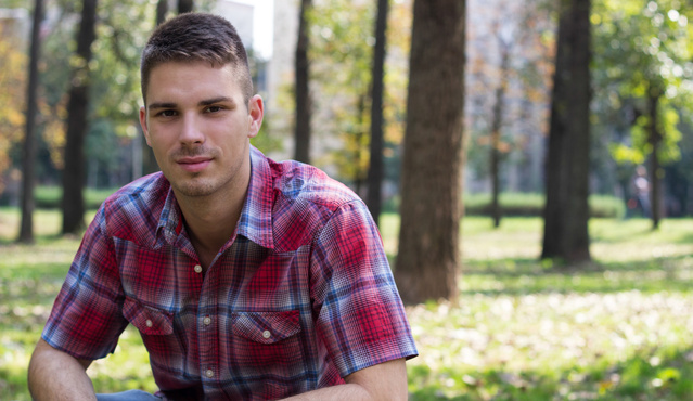 A young white man wearing a red plaid shirt looks at the camera while sitting in front of a wooded area